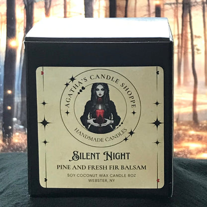 A soy candle with a label featuring a woman's face and the text 'Silent Night Pine and Fresh Fir Balsam'. The candle is presented in a forest setting with blurred lights and trees in the background.