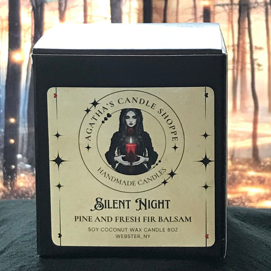 A soy candle with a label featuring a woman's face and the text 'Silent Night Pine and Fresh Fir Balsam'. The candle is presented in a forest setting with blurred lights and trees in the background.