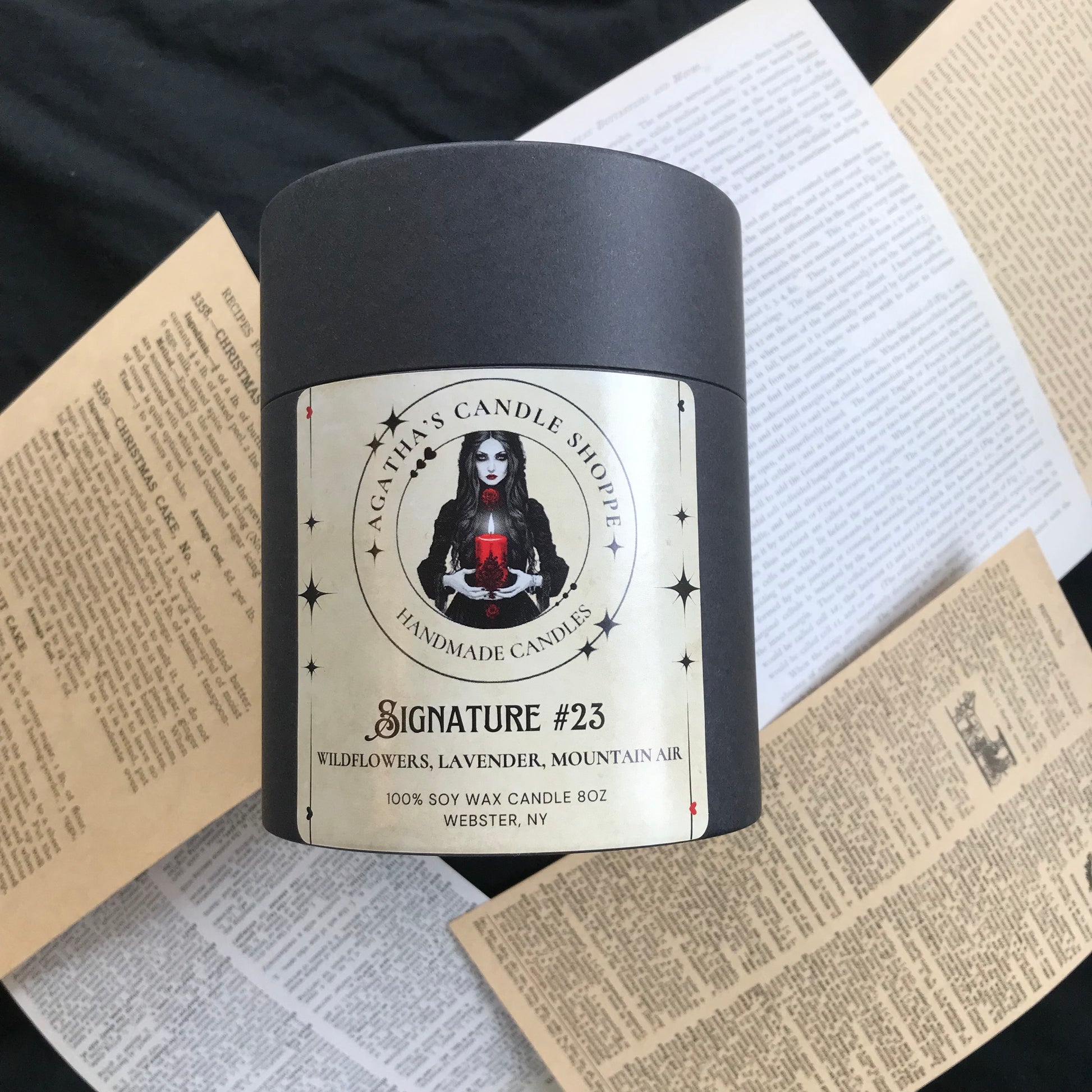 A soy candle in its box, with pages from books underneath telling the story of mystery and love