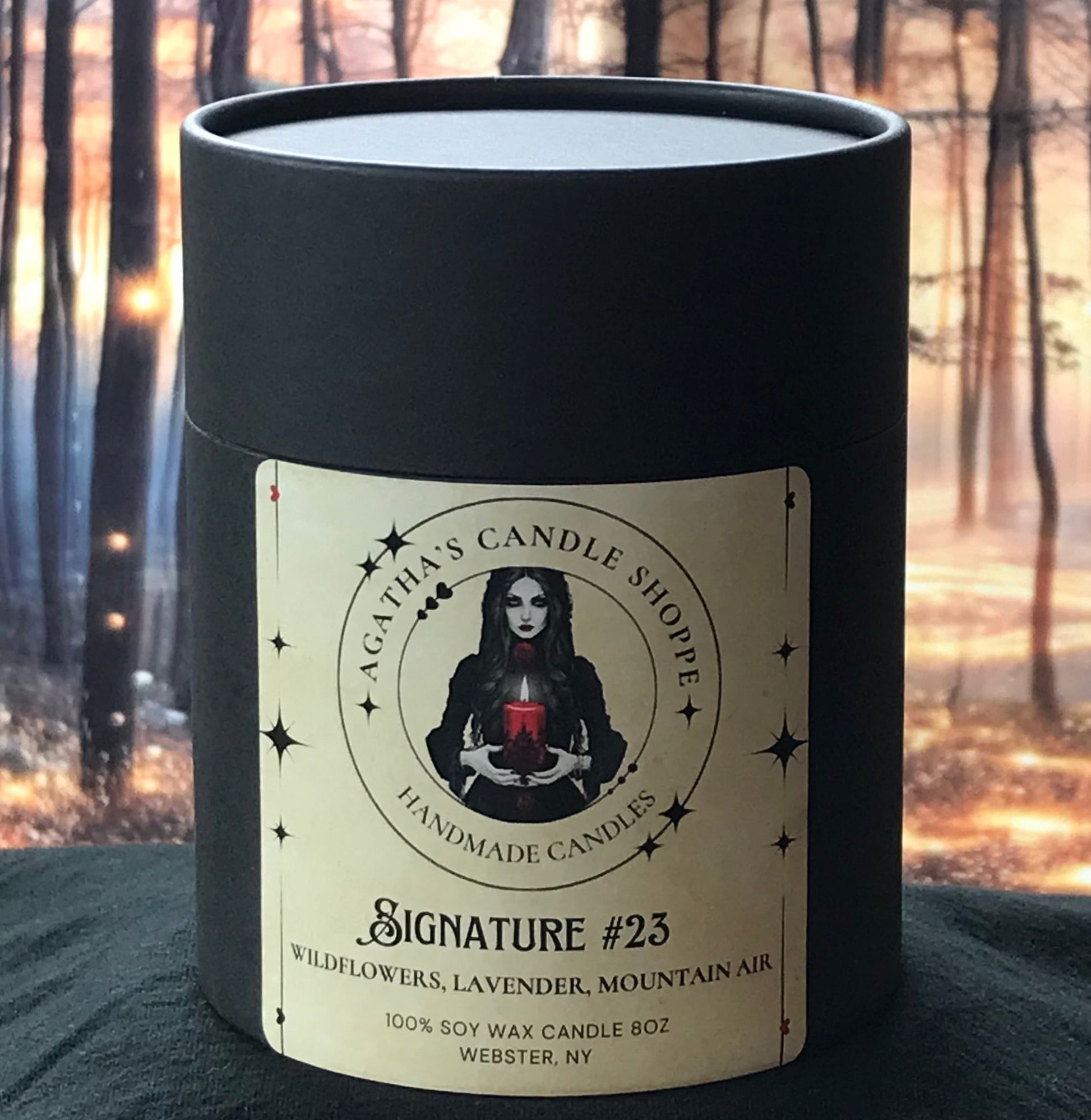 A soy candle labeled 'Agatha's Signature 23'  with a backdrop of a forest and magical, lights.