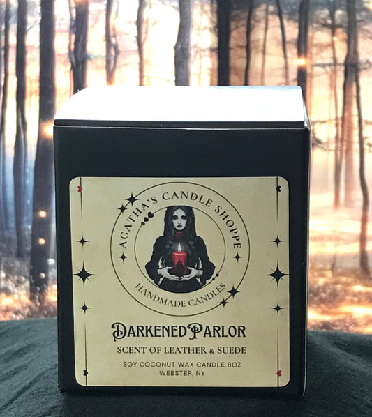 A black soy candle with a label featuring a woman's face and the text 'Darkened Parlor, Scent of Leather & Suede, 8 oz Coconut Soy Candle'. The candle is presented in a mystical nature-inspired setting with trees and magical lights in the background.